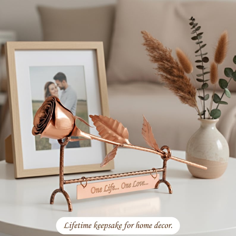 May include: A copper-coloured metal rose sculpture resting on a matching stand with a rectangular plaque engraved with the text One Life... One Love... The rose features a single bloom and two leaves on a stem, supported by a textured metal frame.