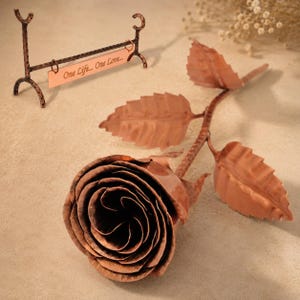 Op de afbeelding: Een koperkleurige metalen roos met een gestructureerde steel en bladeren, vergezeld van een bijpassende metalen standaard met een rechthoekig plaatje gegraveerd met de tekst One Life... One Love... in een cursief lettertype.