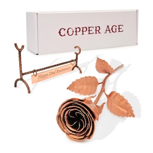 May include: A copper rose with detailed petals and leaves, beside a copper sign that reads "Happy 22nd Anniversary". The rose is on a white surface, with a box that says "COPPER AGE".