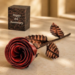 May include: A metallic red rose with a long stem and copper-coloured leaves, accompanied by a dark, textured rectangular block featuring the engraved text "Happy 8th Anniversary" in a light-coloured font on its front face.