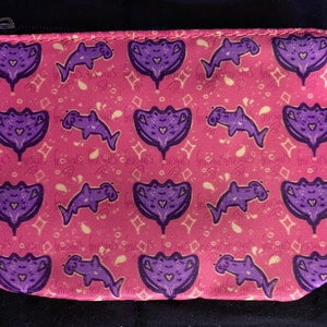 May include: Pink zippered pouch with a repeating pattern of purple dolphins and stylized purple manta rays. The design includes small white diamond and paisley shapes. The pouch has a silver zipper.