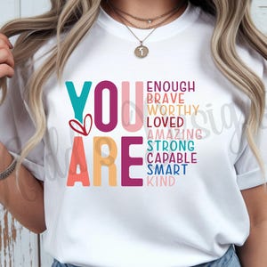 May include: A white short-sleeved t-shirt featuring the large text YOU ARE in teal, maroon, coral, and yellow. To the right, a list of words in various colors includes ENOUGH, BRAVE, WORTHY, LOVED, AMAZING, STRONG, CAPABLE, SMART, and KIND.