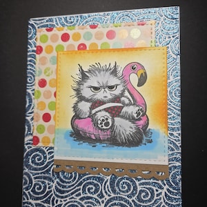 May include: A handmade greeting card featuring a grumpy gray cat floating on a pink flamingo pool float. The card has a blue glitter wave pattern border, a polka dot background, and a brown scalloped edge.