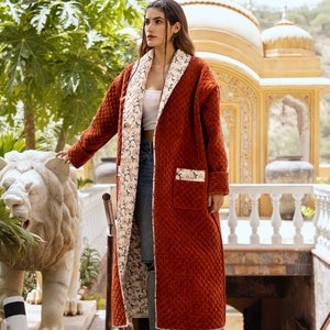 May include: A long, rust-coloured quilted coat with a floral print lining. The coat has a shawl collar and a single patch pocket on the left side. The model is wearing the coat open, revealing the floral lining and a pair of blue jeans.