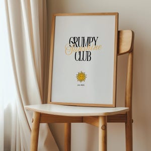 May include: A framed print with the text "GRUMPY SUNSHINE CLUB est. 2022" in black and gold lettering. A yellow sun with a smiling face is below the text.