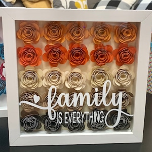 May include: A white shadow box with a glass front, decorated with paper flowers in shades of orange, beige, and gray. The flowers are arranged in rows, and the text "family is everything" is written in white on the front of the box.