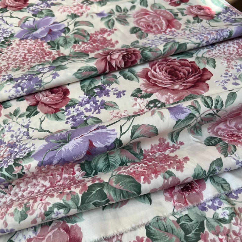 Pode incluir: Um tecido branco com um padr&atilde;o floral com rosas rosa e roxas, folhas verdes e pequenas flores roxas.