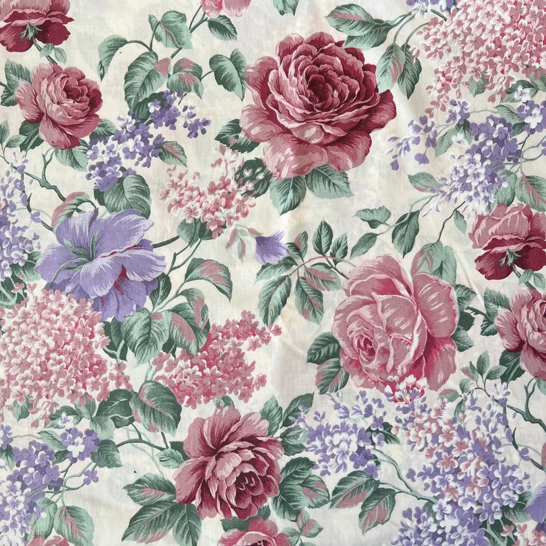 Pode incluir: Um tecido floral com um padr&atilde;o repetido de rosas cor-de-rosa e flores de lil&aacute;s roxas sobre um fundo creme.
