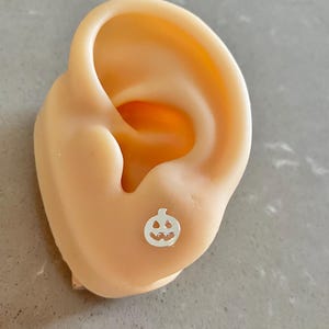 Puede incluir: Un pendiente de botón de plata con forma de calabaza. El pendiente es una pequeña y detallada representación de una calabaza de Halloween. Se muestra en un modelo de oreja color carne, sobre un fondo gris.