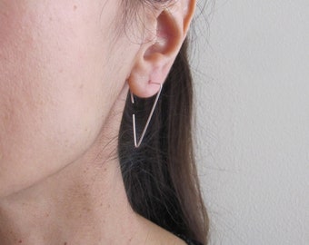 May include: A pair of silver triangle shaped hoop earrings. The earrings are worn in the left ear of a person with long dark hair.