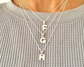 以下が含まれることがあります： イニシャルペンダントが付いたシルバーネックレス3本。ペンダントはF、G、Hの文字。ネックレスは重ねられています。