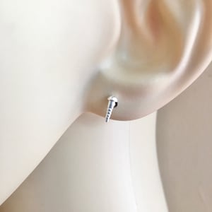 Könnte beinhalten: Nahaufnahme eines silbernen Ohrsteckers in Form eines kleinen, spitzen Kegels. Der Ohrstecker wird im linken Ohrläppchen einer Puppe getragen.