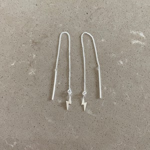 May include: A pair of silver threader earrings. Each earring has a fine chain, a small lightning bolt charm, and a straight bar at the top. The earrings are shown against a neutral grey background.