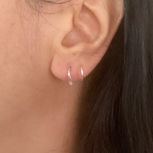 Puede incluir: Primer plano de una oreja con dos pequeños pendientes de aro de plata. Los pendientes son simples y delicados, y son una opción popular para el uso diario.