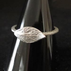Può includere: Un anello d'argento con un design a foglia dettagliato. La foglia è il punto focale, con venature intricate. La fascia ha un aspetto martellato e strutturato. L'anello è esposto su un supporto nero, evidenziando la sua lucentezza metallica.