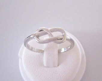 May include: A silver infinity knot ring. The ring is on a white display stand.