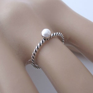 May include: A silver-colored ring with a twisted band and a small, round bead at the top. The ring is shown on a person's finger, highlighting its delicate design and minimalist style. The ring is a piece of jewelry.