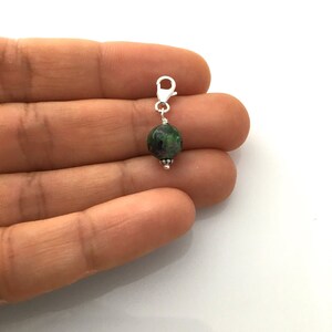 May include: A silver charm with a round green and pink gemstone bead attached to a silver lobster clasp.
