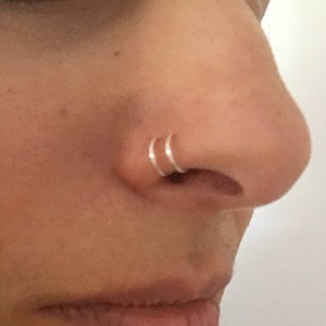 Puede incluir: Primer plano de la nariz de una persona con dos anillos de nariz plateados. Los anillos son finos y delicados, y se llevan en la fosa nasal.