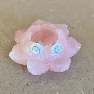 May include: Stud earrings with a spiral design, displayed on a pink, flower-shaped dish. The earrings feature a light blue centre with a gold spiral. The dish is a soft pink colour and is placed on a neutral surface.