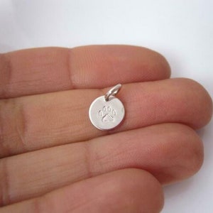 May include: Silver charm featuring a paw print design. The charm is round and has a loop for attaching to a necklace or bracelet.