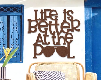 Puede incluir: Un letrero de madera marrón con el texto "Life is better at the pool" con un par de gafas de sol en lugar de la "O" de pool. El letrero está colgado en una pared blanca junto a una puerta azul. Una silla de mimbre con un cojín a cuadros se encuentra frente al letrero.