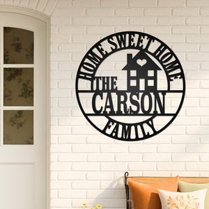 Puede incluir: Letrero de metal negro "Home Sweet Home" con una silueta de casa y el apellido "The Carson Family". El letrero está montado en una pared de ladrillo blanco cerca de una puerta blanca y un banco con cojines.