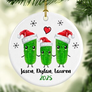 May include: A white ceramic Christmas ornament featuring three cartoon pickles wearing Santa hats. The ornament has the names "Jason, Dylan, Lauren" and the year "2025" printed on it, along with snowflakes and a red heart.