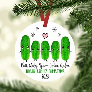 May include: A white round Christmas ornament with five green pickle characters holding hands. The pickles have smiling faces and small arms and legs. The text "Brett, Wesley, Spencer, Jackson, Andrea HOGAN FAMILY CHRISTMAS 2023" is printed below the pickles.