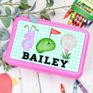 Puede incluir: Una fiambrera rectangular de plástico rosa con un patrón de cuadros vichy en verde claro y blanco. La tapa muestra una bolsa de golf, un hoyo de golf verde con una bandera roja y una pelota de golf sobre un tee. El nombre BAILEY está en negro.