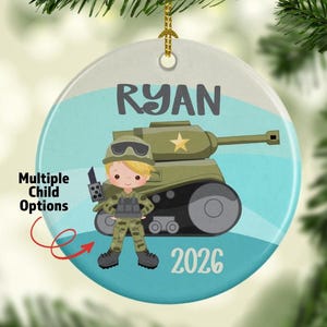 May include: A round ceramic ornament featuring a cartoon illustration of a child in camouflage military gear standing in front of a green tank. The name RYAN is printed in dark gray at the top, and the year 2026 is printed at the bottom.