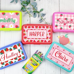 May include: Five rectangular plastic pencil boxes in lime green, pink, red, blue, and teal, each featuring a personalized name label and strawberry-themed patterns. A box of 24 Crayola Ultra-Clean Washable Crayons and an apple-shaped cutout are included.