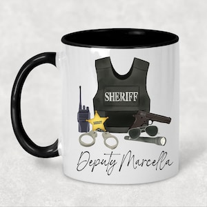 May include: A white ceramic mug with a black handle and rim, featuring a graphic of a black sheriff vest, a walkie-talkie, a gold star badge, handcuffs, a handgun, sunglasses, and a flashlight, with the text Deputy Marcella printed below.