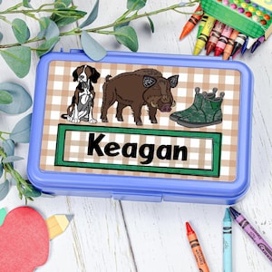 May include: A rectangular blue plastic pencil case featuring a beige and white checkered background. The lid displays a graphic of a black and white dog, a brown wild boar, and green camouflage boots, with the name Keagan printed in black text.