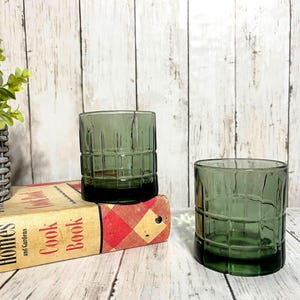Könnte beinhalten: Zwei grün getönte Trinkgläser mit quadratischem Muster. Die Gläser sind zylindrisch mit dickem Boden. Sie stehen auf einem Vintage-Kochbuch mit den Worten "Better Homes and Gardens Cook Book". Im Hintergrund befindet sich eine kleine Topfpflanze.