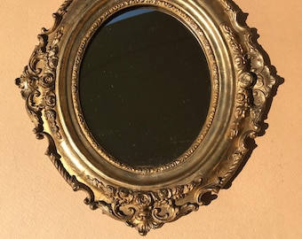 May include: A gold oval mirror with an ornate, detailed frame. The frame has a raised, textured design with floral motifs.