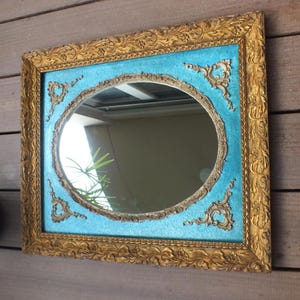 Könnte beinhalten: Ein quadratischer Spiegel mit einem verzierten goldenen Rahmen und einem türkisblauen Hintergrund. Der Spiegel hat eine runde, ovalförmige reflektierende Oberfläche.