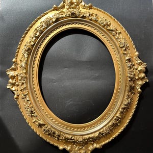 Könnte beinhalten: Ein ovaler Bilderrahmen mit metallisch goldener Oberfläche. Der Rahmen weist aufwendige Reliefschnitzereien mit Blumenmustern, geschwungenem Blattwerk und dekorativen Perlenverzierungen entlang der Innenkante auf, die ein strukturiertes Aussehen verleihen.