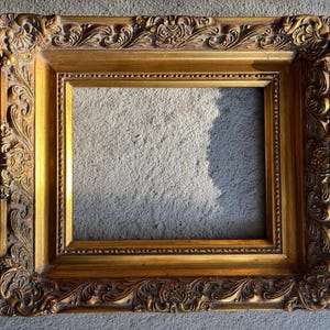 Könnte beinhalten: Ein rechteckiger Bilderrahmen mit metallisch goldener Oberfläche. Der Rahmen weist auf seinem breiten Rand aufwendige, erhabene Schnörkel- und Blumenreliefmuster auf, mit einem glatten, vertieften Innenrand um die zentrale Öffnung.