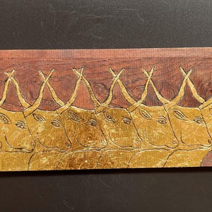 May include: A rectangular wooden plaque featuring a carved and gold-leafed relief of a row of cattle. The natural dark brown wood grain is visible at the top, while the lower section displays a series of stylized bull heads with long horns.