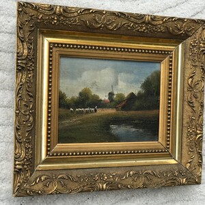 Op de afbeelding: Een ingelijst olieverfschilderij dat een landelijk landschap met een windmolen, een boerderij en een groep schapen op de voorgrond toont. Het schilderij is ingelijst in een gouden lijst met sierlijke details.