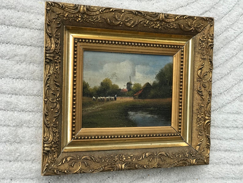 Op de afbeelding: Een ingelijst olieverfschilderij dat een landelijk landschap met een windmolen, een boerderij en een groep schapen op de voorgrond toont. Het schilderij is ingelijst in een gouden lijst met sierlijke details.