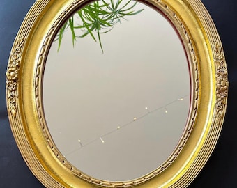 May include: Oval gold-framed mirror with ornate floral detailing. The mirror's frame has a textured, ridged design and a rich, golden hue. The mirror reflects a room with a plant and string lights, creating a vintage aesthetic.