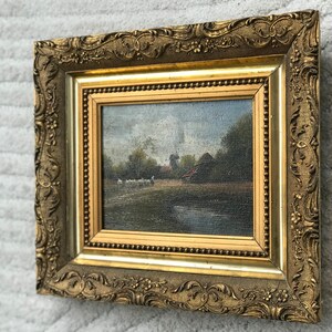 Op de afbeelding: Een ingelijst olieverfschilderij dat een landelijk landschap met een windmolen, een boerderij en een groep schapen op de voorgrond toont. De lijst is versierd en goudkleurig.