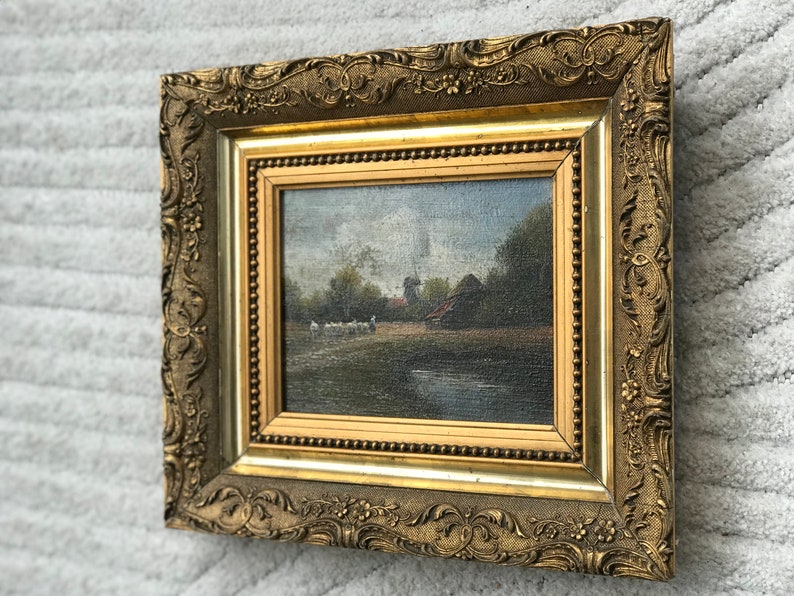 Op de afbeelding: Een ingelijst olieverfschilderij dat een landelijk landschap met een windmolen, een boerderij en een groep schapen op de voorgrond toont. De lijst is versierd en goudkleurig.