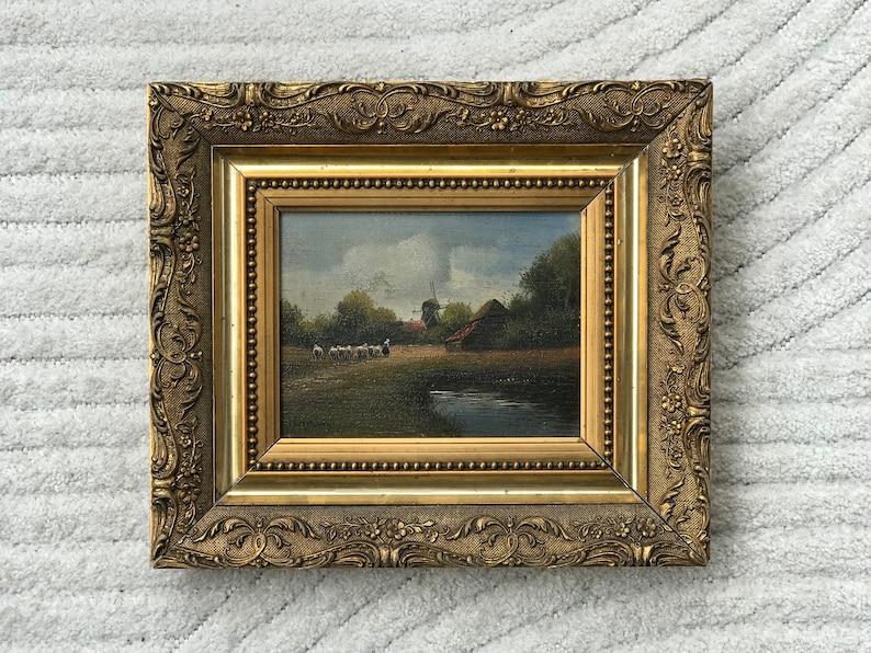 Op de afbeelding: Een ingelijst olieverfschilderij dat een landelijk landschap met een windmolen, een boerderij en een kudde schapen afbeeldt. Het schilderij is ingelijst in een gouden lijst met sierlijke details.