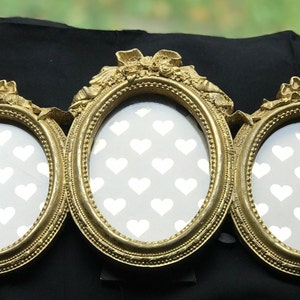 Puede incluir: Un marco de fotos triple en tono dorado con detalles ornamentados y aberturas ovaladas. Cada abertura presenta un patrón de corazón blanco sobre un fondo gris claro.