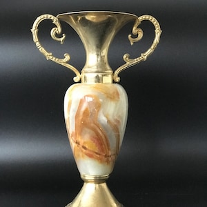 May include: A gold and white marble vase with ornate gold handles. The vase has a classic, elegant design.