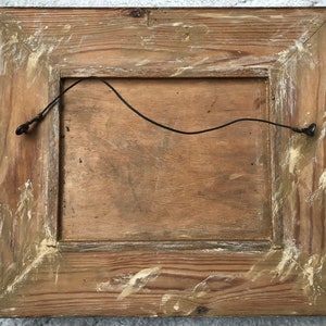 Op de afbeelding: De achterkant van een houten fotolijst met een draadhanger eraan bevestigd. De lijst is verweerd en heeft een lichtbruine afwerking.