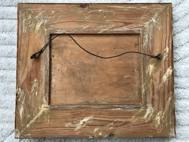 Op de afbeelding: De achterkant van een houten fotolijst met een draadhanger eraan bevestigd. De lijst is verweerd en heeft een lichtbruine afwerking.